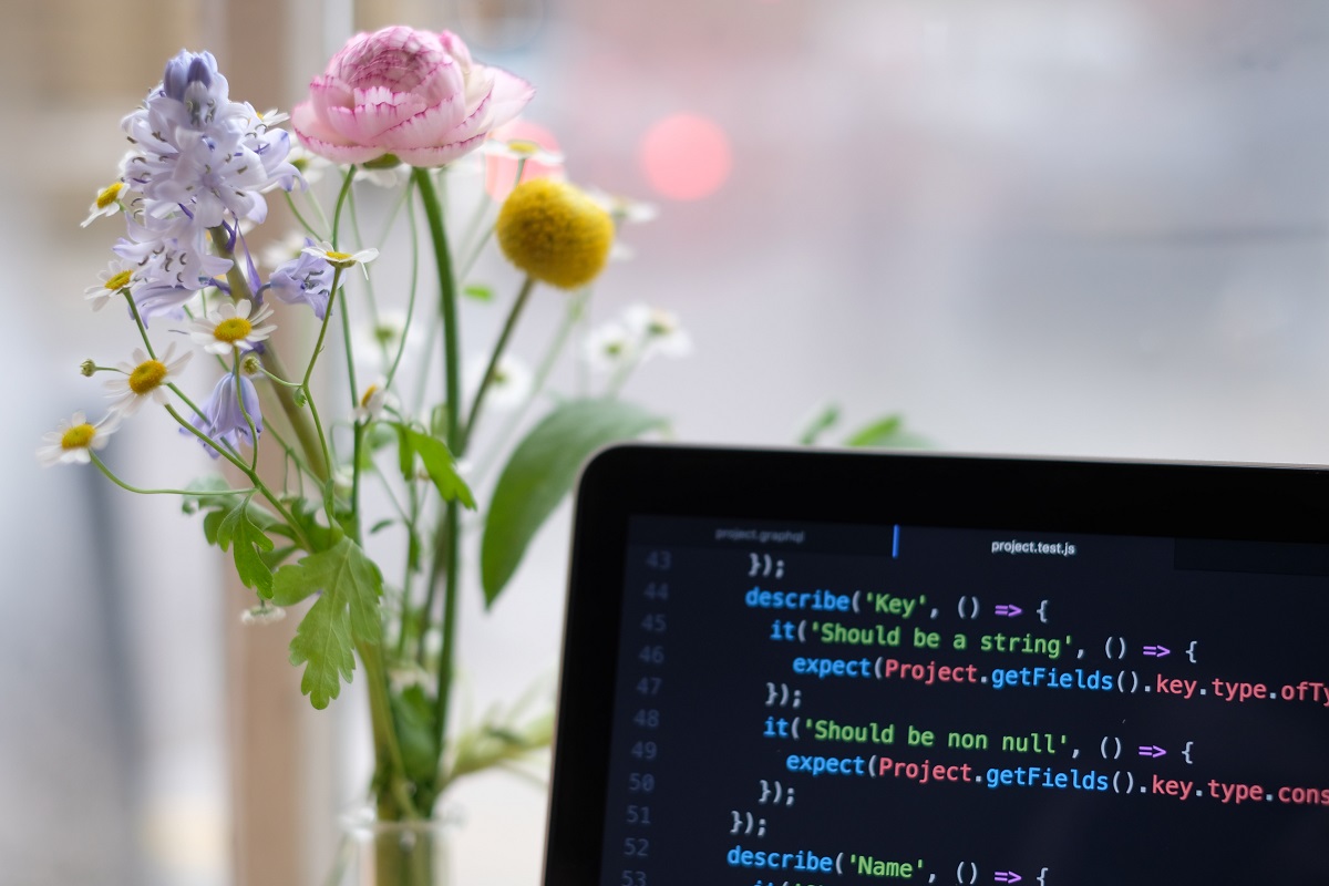 Featured image for the post "JaneBuzJane's Userscripts." The corner of a laptop running pastel code is visible in the lower right corner, alongside a bright bunch of flowers in a vase on the left side.